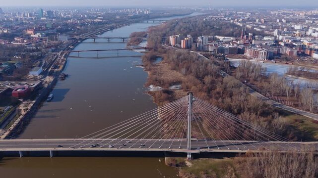 Aerial view shows Swietokrzyski Bridge in Warsaw as cars move, single pylon and fan cables leading to older bridges, promenade, ice floes, long shadows in daylight.