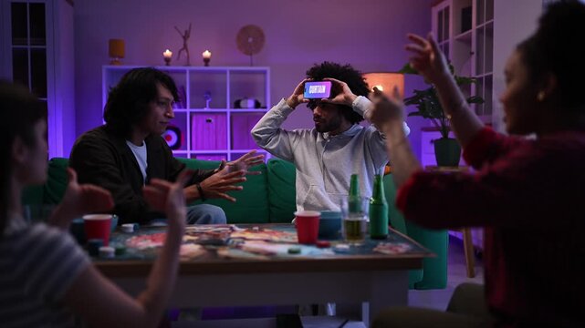 Group of diverse friends playing a word guessing game with a smartphone on a man's forehead at a party in a neon lit room