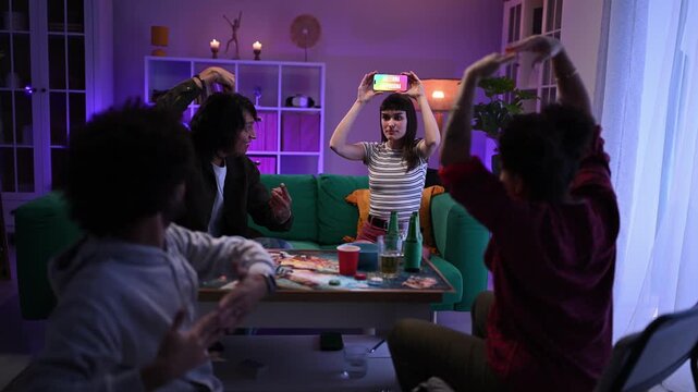 Diverse group of young friends laughing and having fun together while playing a word guessing game on a smartphone during a home party