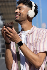 Indian man standing on sunny street, smiling, wearing headphones, smartwatch, bag, holding wrapper © wavebreak3