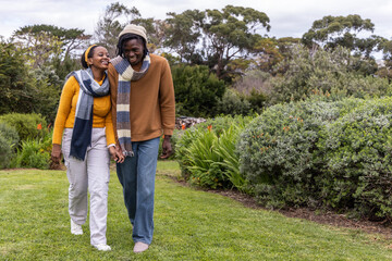African American couple walking hand in hand on grassy lawn at park, wearing sweaters and scarves