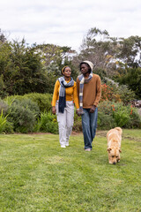 African American couple holding hands walking in park wearing sweaters and scarves with tan dog