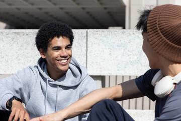 Diverse male teenagers sitting on campus steps, in hoodie, chatting with headset and smartwatch © wavebreak3