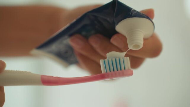 Closeup toothpaste squeezed onto toothbrush hotel bathroom, guest hand applies mint paste onto pink bristles, soft natural light, shallow depth of field, minimal aesthetic, intimate hygiene routine,