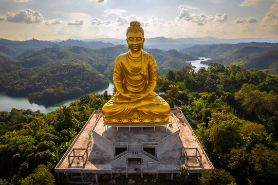 Breathtaking aerial panorama of the large golden Phra Phuttha Lokanat Satsada meditation Buddha statue at Wat Wang Wiwekaram overlooking winding river, lush forest, and layered mountain landscape in S