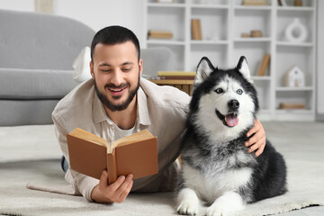 Handsome young man lying on floor and reading book with cute husky dog at home © Pixel-Shot