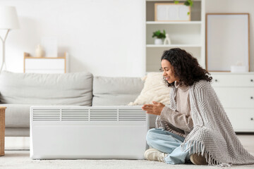 Obraz premium Young African-American woman with plaid warming near electric convector heater at home