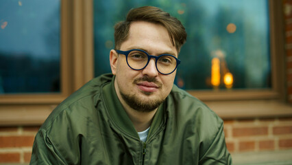 Obraz premium Portrait of a handsome young bearded caucasian man in blue glasses and a green bomber jacket relaxing outdoors near a brick wall.