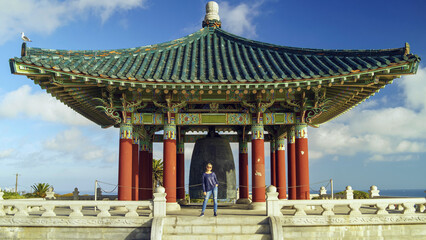 Female tourist stands by the famous Korean Bell of Friendship pavilion in Angel's Gate Park under a blue sky with white clouds. © AncientAftertone
