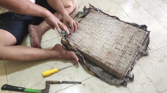 Close up of a craftsman removing old upholstery staples from a woven bamboo chair seat using pincers during furniture restoration on a tiled floor