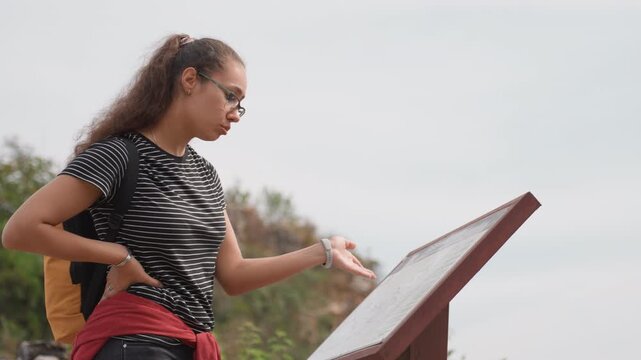 Young woman traveler studies map panel and considers route, hands on hip, yellow backpack visible and red jacket tied at waist, thoughtful posture against rocky overlook, cloudy sky, planning detour