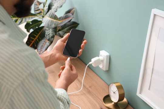 Young man connecting charging cable to mobile phone at home, closeup