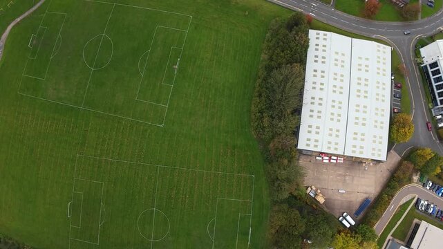 Aerial View Football Pitches Leeds, UK Showing Whiteroof Warehouse Beside Green Community Fields, Marked Soccer Lines, Parking Yard, Tree Belt And Adjacent Residential Road. Calm Overcast Light