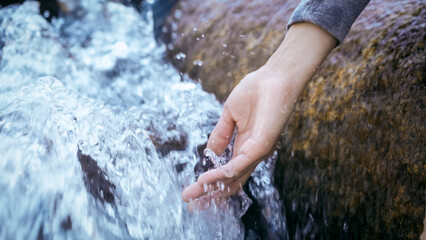 Close-up of a human hand reaching into a fast-flowing clear mountain stream beside a mossy rock,...