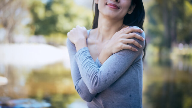 Young woman outdoors hugging herself, wet gray shirt, calm smile, soft natural light and blurred park background, suggesting freshness, comfort, or wellness concept.