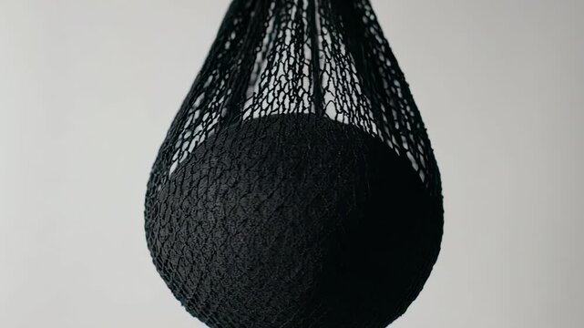 Mesh Ball Bag. Football Training Equipment Mockup (Functional & Educational). Grassroots Training. An empty black mesh ball-carrying bag hanging from a hook, swaying slightly, set against a white wall