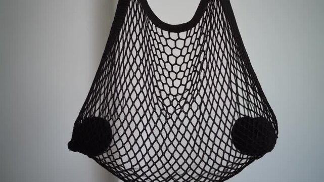 Mesh Ball Bag. Football Training Equipment Mockup (Functional & Educational). Grassroots Training. An empty black mesh ball-carrying bag hanging from a hook, swaying slightly, set against a white wall