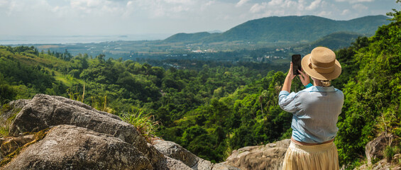 Young woman in straw hat taking photo of tropical valley and mountains from rocky viewpoint in Thailand with lush jungle, distant sea, and wide sky creating natural copy space for travel themes © StockMediaSeller