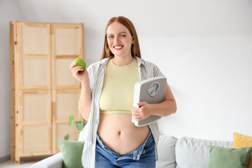 Young happy overweight woman in tight jeans with fresh apple and scales at home