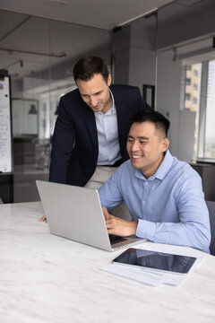 Supportive office mentoring. Smiling mid aged businessman mentor stand beside young male intern apprentice sitting by notebook offer guidance share insight give positive feedback on project solution