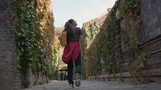 Back view woman walking ivy alley, yellow backpack and red jacket tied at waist, cobblestone path framed by climbing vines, sunlight filtering through leaves, cinematic tracking shot of casual