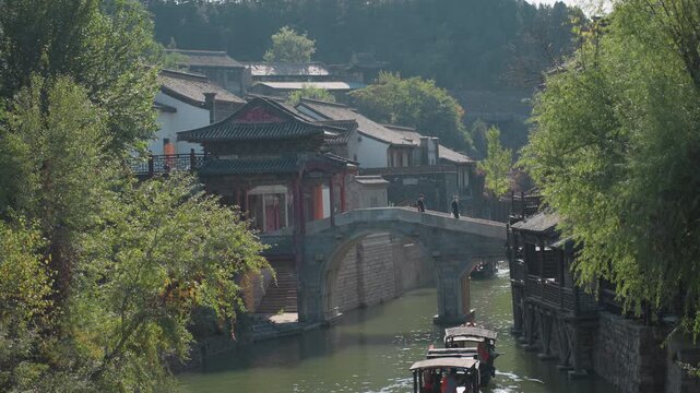 Sunlit canal stone bridge red pavilion nestled among willow trees, wooden boatman guides slow skiff, morning light sparkles on water, tranquil historic village atmosphere, heritage architecture