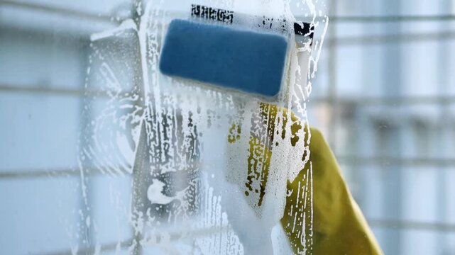 Close up of a person wearing yellow gloves using a blue sponge to scrub and clean a window with soapy water, showing the movement of washing and leaving a clean surface in its wake