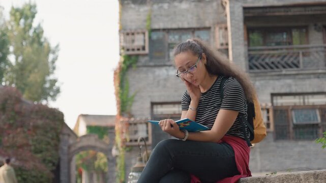 Student journaling on stone steps with pen and glasses focused writing in notebook backpack at side historic courtyard backdrop ivy covered walls concentrated learning and creative notes