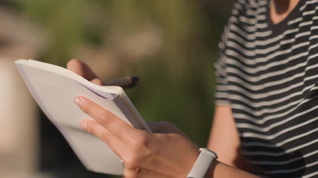 White woman taking notes in park, close view of hands with notebook and pen, striped shirt and smartwatch, focused planning and schedule checking, soft morning sunlight and greenery blur
