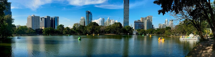 Fototapeta premium Colorful swan paddle boats glide across the calm lake at Lumpini Park in Bangkok, framed by lush trees and a modern skyline under clear blue skies on a bright afternoon.