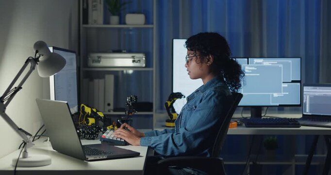 Young African American female programmer coding AI software on multiple screens in night office. Network security, cybersecurity, machine learning, working late, overtime, robotics automation concept.