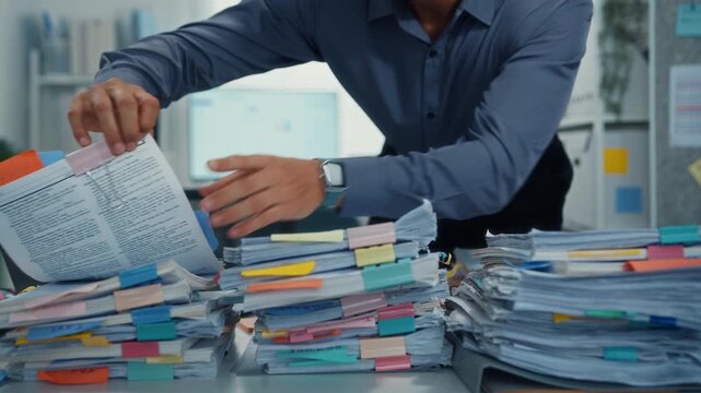 Overworked young Latin businessman reading document in large stack of paperwork files at office desk. Tax audit deadline, accounting stress, corporate burnout, bookkeeping, bureaucracy concept.