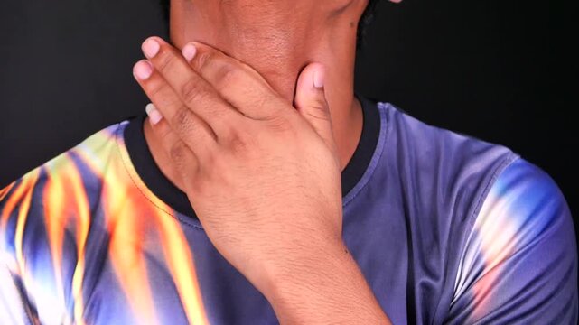 Man rubbing sore throat and massaging painful neck area against dark background in close up cinematic footage