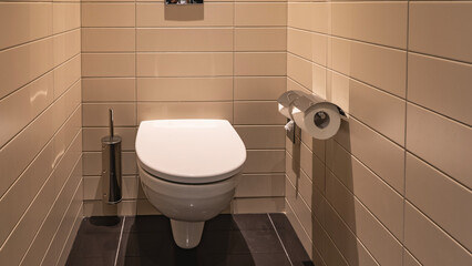 Modern lavatory. Beige tiled walls and floor. White porcelain toilet bowl. A brush and toilet paper rolls.