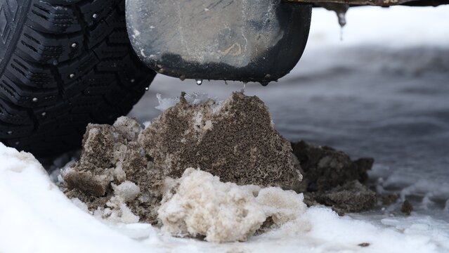 Close-up of dirty slush and ice buildup on the ground next to a car tire and mudflap during winter.