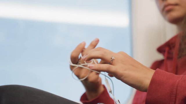 Close up frantic hands holding tangled earbuds against window, city skyline beyond glass, passenger fumbles cable and adjusts plug while checking phone, palpable hurry and small annoyances of commute.
