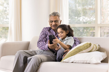 Older 70s man spend time on internet with his little 6s great-grandson, sit on sofa, looking at...