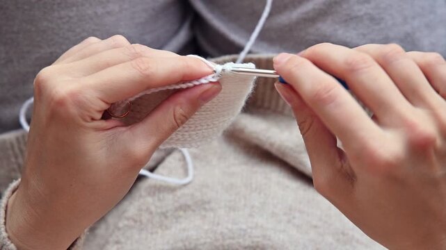 Female artisan with light skin using a blue crochet hook to create a white knitted piece while seated on a couch with a beige sweater and soft fabric background