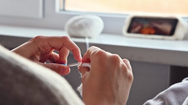 Female hands skillfully crochet with a blue hook, manipulating white yarn while a smartphone displays a tutorial video in a cozy indoor setting