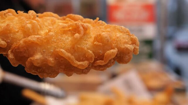 Extreme close-up of golden crispy batter on a piece of deep-fried fish fillet, symbolizing classic British fish and chips or fast food street fare.