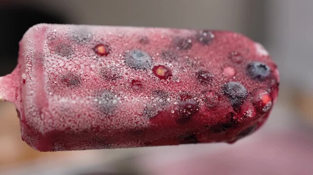 Macro shot of frozen mixed berry ice pop on a wooden stick, showing frosty texture and whole fruit pieces, ready to be enjoyed as a refreshing summer treat.