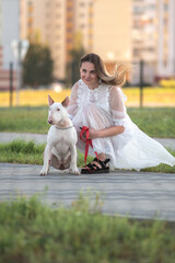 Caucasian woman in white dress kneels beside a white bull terrier dog on a paved path with green grass and urban buildings in the background during daylight
