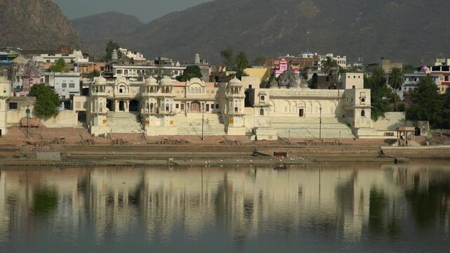 Wide shot of Pushkar Lake with historic palace and ghats reflected in calm water, Rajasthan, India. Traditional Hindu architecture and sacred waterfront in morning light.