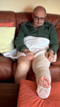 Relaxed elderly man with a bandaged leg resting on the sofa in his home