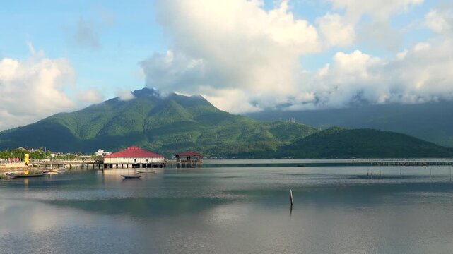 Quiet morning at Lap An Lagoon featuring a red-roofed restaurant over still water against lush, cloud-touched green mountain peaks.