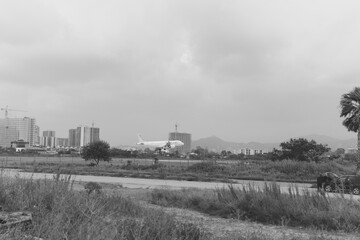 Grayscale landscape with distant city skyline, open field, scattered shrubs, and a grazing horse on the right under a cloudy sky, capturing a calm rural mood near urban development.