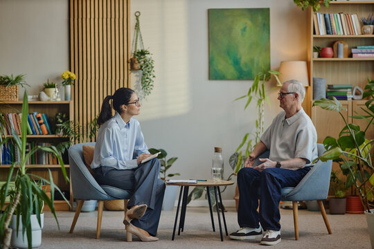 Middle aged Caucasian woman conducting therapy session with senior Caucasian man sitting across in modern office setting, both engaged in conversation, psychology concept