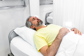 Obraz premium Mature male patient lying in hospital bed in clinical ward. Sick or recovering man resting on white pillow under blanket. Healthcare and medicine concept of patient care and hospital stay recovery.