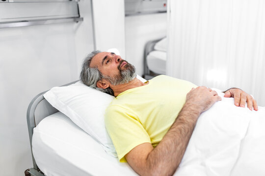 Mature male patient lying in hospital bed in clinical ward. Sick or recovering man resting on white pillow under blanket. Healthcare and medicine concept of patient care and hospital stay recovery.