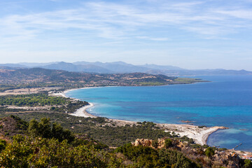 Fototapeta premium Capo Comino Faro Vecchio lighthouse overlooking Mediterranean coastline. Sandy beach and turquoise waters in Sardinia, Italy. Scenic view of rocky shoreline and distant mountains.